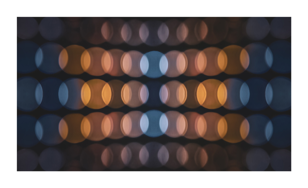The photo is a pattern of blurred, out-of-focus circles arranged in rows and columns. The circles alternate in colour between warm, orange-toned hues and cooler, blue-toned hues. The background is dark, and emphasizes the glowing effect of the circles.