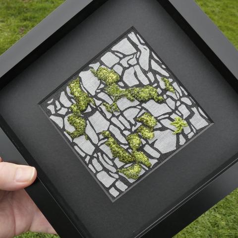 Lino cut print on fabric depicting a dry stone wall (black on mottled grey fabric). Stitching has been added in shades of green depicting moss growing on the wall. Framed in black and held in the artist's hand for scale