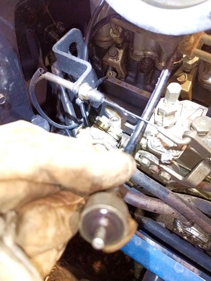 Photo of a tractor engine. In the front, out of focus, a gloved hand holds up a threaded fitting and a keyed shaft that goes into a dark gray painted tube. Behind, in focus and with a headlamp shining on it, the fitting on the top of the injector pump where the shaft goes.