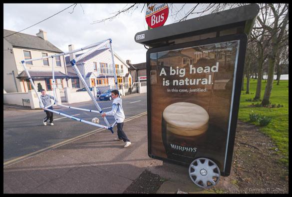 Street photograph of two young boys carrying a blue and white portable football goal across a road in Cork, Ireland. In the foreground is a Bus Éireann bus shelter with a Murphy's Irish Stout advertisement featuring a pint glass with creamy head and the text "Enjoy MURPHY'S responsibly", "A big head is natural. In this case, justified." and the Murphy's logo. A red "Bus Éireann Bus" sign sits atop the shelter marked "2377". Background shows traditional Irish houses, a blue car, and a green park with bare trees.