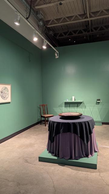 A contemporary art gallery with painted sage green walls.

The centerpiece of the installation is a circular table with teal and lilac tablecloths draped to the floor, elevated by a low plinth. There is a spinning circular lazy Susan at the center of the table made of a lacquered wood featuring an inlaid woodcut.

Mirrors on the far wall cast crystal-like reflections. There is a wooden dining chair in the corner and framed artworks on the walls.