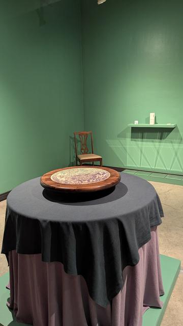 A contemporary art gallery with painted sage green walls.

The centerpiece of the installation is a circular table with teal and lilac tablecloths draped to the floor, elevated by a low plinth. There is a spinning circular lazy Susan at the center of the table made of a lacquered wood featuring an inlaid woodcut.

Mirrors on the far wall cast crystal-like reflections. There is a wooden dining chair in the corner and framed artworks on the walls.