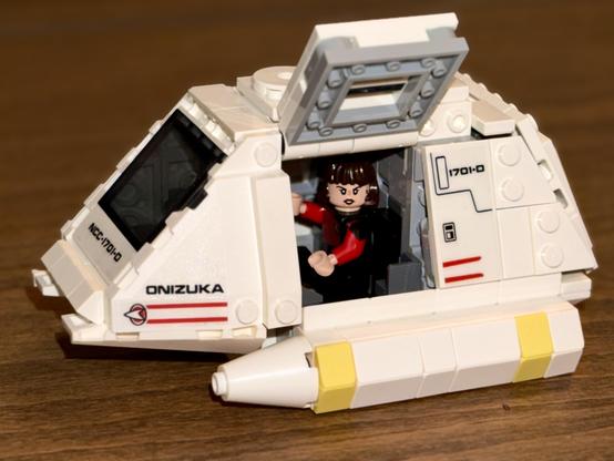 A shallow DOF close view of a Lego version of a Star Trek the Next Generation type 15 shuttle craft with the gull-wing door open revealing a small female figure at the controls wearing a red and black Star Trek TNG uniform