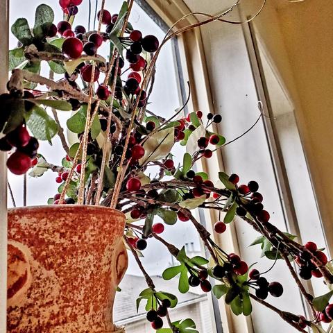 An indoor close-up shot of a decorative arrangement in a terra-cotta colored pot situated near a window. The arrangement features several sprigs, branches, and stems covered in red and dark-colored, glossy berries. Some green leaves and gold-colored, glittery sprigs are also part of the arrangement. A portion of the window frame and an exterior view are visible in the background.