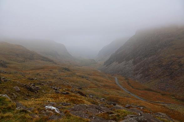 This image captures a dramatic and atmospheric landscape of a mountainous valley shrouded in mist. The scene is dominated by rolling hills and rugged, rocky slopes that stretch into the distance, their earthy brown and green hues suggesting autumnal vegetation. A winding road cuts through the valley floor, with a single car visible, disappearing into the foggy horizon and adding a sense of mystery and depth to the composition.

The mist obscures the upper portions of the mountains, blending seamlessly with the overcast sky and creating a soft, ethereal quality. The overall mood is one of tranquillity and solitude, with the muted colours and fog evoking a sense of calm and introspection.