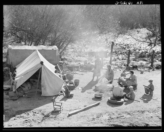 The black and white photograph captures the life of two families, comprising fifteen people from Chickasaw, Oklahoma. They are shown camped by the roadside near Santa Maria, California during a time when they were heading towards the pea harvest but unable to proceed due to lack of funds for purchasing a trailer's license. The image depicts them in an outdoor setting with makeshift tents and various camping paraphernalia spread out on the ground around them. Several individuals are visible; some seated or squatting while others stand, engaging in activities such as cooking over a campfire, carrying containers, and handling supplies. There is a sense of hardship conveyed by their surroundings and actions, highlighting their struggle for sustenance during what appears to be challenging times economically. The photograph underscores the harsh realities faced by migrant families seeking work opportunities across different regions.