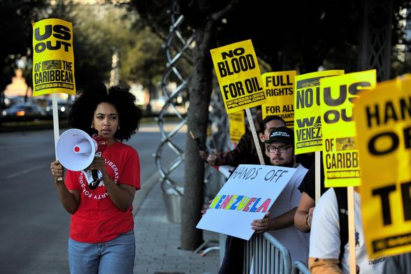 On the left side of the frame stands a Black woman in a red Party for Socialism & Liberation shirt holding a megaphone and a sign that says "US out of the Caribbean". She's in-between chanting out and is looking towards the crowd joining the protest on the other side of the police barricade. The crowd is filled with similar signs denouncing the US' escalation against Venezuela.