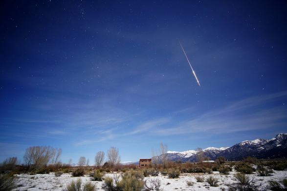 A photograph of the night sky under a waning gibbous moon. The moon is bright enough that the sky is colored blue, and there are some stars visible. A meteor streaks diagonally right of center, aimed toward the bottom right. The tail is thin, and gets thicker at the terminus. There are some cirrus clouds, thicker on the horizon. The snow-capped peaks of the Rocky Mountains stretch from center to right on the horizon. Most of the foreground is snow-covered, with scattered chamisa bushes, cottonwood and aspens bare branches sticking up. A single two-story strawbale house sits center of the frame.