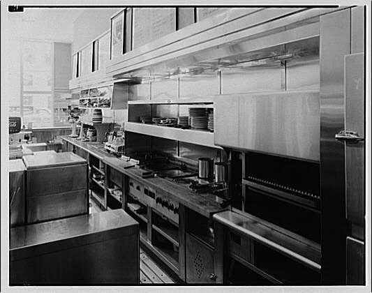 The image displays a black and white photograph of an industrial kitchen setting, likely from the early to mid-20th century. The space is equipped with stainless steel appliances such as ovens, hoods, storage cabinets, and preparation tables. There are stacks of plates placed on shelves above the work surfaces, indicating readiness for serving food.
The layout suggests a clean and efficient workflow designed for high-volume meal preparation. A large oven dominates one side of the kitchen, while other equipment is organized along the walls in an orderly fashion. The absence of people in the image emphasizes the focus on the environment rather than individual workers or their actions within this setting.
This photograph captures elements that reflect a particular historical period's design and technology for commercial food service establishments, which have since evolved with advancements in kitchen appliances and ergonomics to improve safety and functionality for staff.