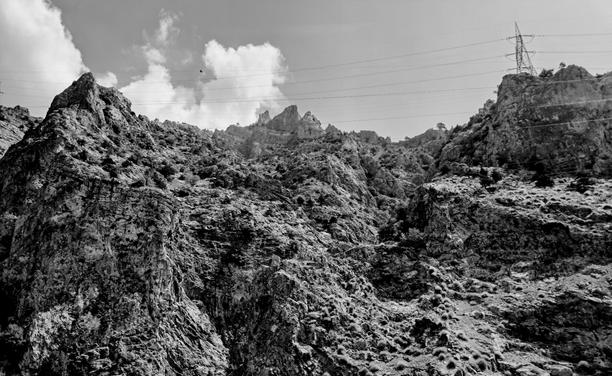 Monochrome picture of rugged steep mountain terrain with deep crevices and sparse scrub vegetation; high-voltage power lines span horizontally across upper portion of frame, connecting to steel lattice tower perched on rocky ridge at right; jagged peaks in center background beneath bright blue sky with fluffy cumulus clouds