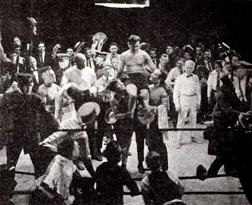 A still from the film. The image is a grainy black and white photograph capturing a boxing match in a crowded arena. The composition is a wide shot, showing a boxing ring in the foreground surrounded by a large crowd.
In the center of the ring, two boxers are the focal point. One boxer, shirtless, is being held up by other men, likely celebrating a victory. He appears to be muscular. The other boxer is likely the defeated one, and is being helped by his corner.
The crowd is densely packed around the ring, with men in suits, hats, and ties. There are also figures in what appear to be uniforms or service attire. The expressions are difficult to discern due to the low resolution, but the overall atmosphere seems excited and celebratory.