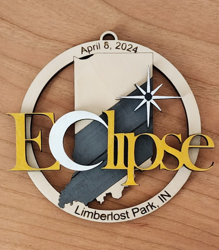 A round ornament with the shape of Indiana in the center, with a large grey area with a single line that follows the path that the 2024 solar eclipse took. There is a compass star over a spot in the upper half of the grey line just above the black line and across the center is the word Eclipse in gold, but the C is a large white sun with the outline of the moon crossing over it. On the circle around it says April 8, 2024 at the top, and Limberlost Park, IN at the bottom.