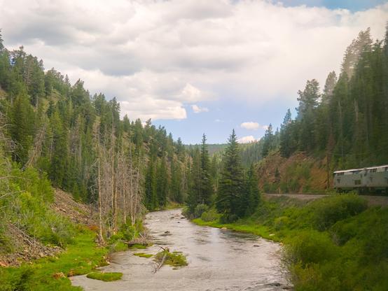 Looking down a medium sized river that appears shallow and a little rocky. The river carves through mountains that are covered in pine forest. A train travels to the right of the river, and its 2 locomotives are visible. It was mostly cloudy.