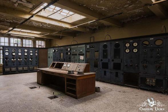 Inside an abandoned power station, a long wooden desk sits in front of old control panels with analog gauges. The panels are labeled "GENERATORS" and "DISTRIBUTION."