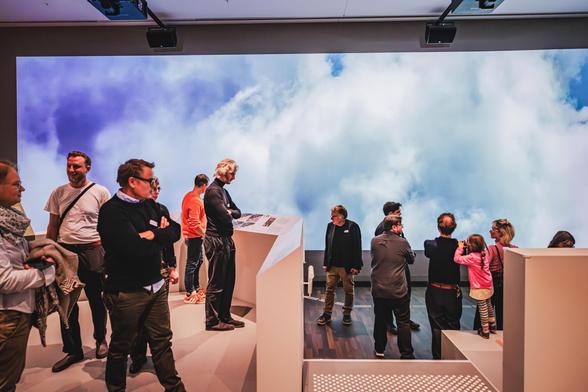Eine Gruppe von Besucher*innen steht in einem Ausstellungsraum des Humboldt Labors. Im Hintergrund ist eine große Wandprojektion mit bewegten Wolken zu sehen. Die Personen betrachten Ausstellungselemente, lesen Informationen oder unterhalten sich. Eine Familie mit einem Kind erkundet gemeinsam einen Bereich der Ausstellung. Die Atmosphäre wirkt lebendig und neugierig. Foto: Stefan Klenke