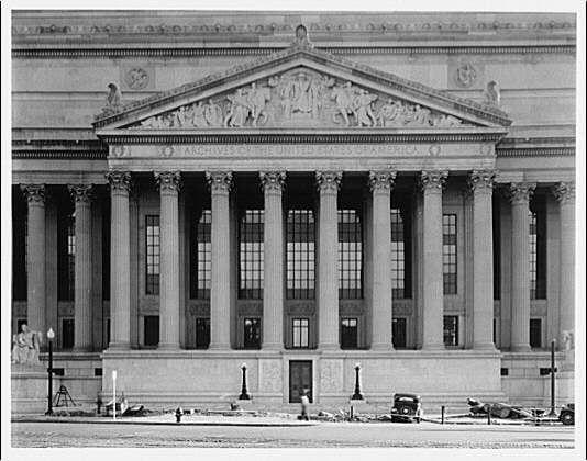 The image depicts the pediment of a large neoclassical building, identified as part of the National Archives. The architecture features prominent columns supporting an elaborate entablature adorned with sculptural decorations and relief work depicting figures in dynamic poses. The structure is set against a background that suggests its urban setting; vehicles are visible on Pennsylvania Avenue below, indicating it's likely located within Washington D.C.

The photograph appears to be taken during the early 20th century through mid-20th century period judging by the style of the cars and street lamps as well as the black-and-white quality of the image. The building exhibits a classical design with a grandiose presence, which is typical for governmental buildings constructed in that era.

Additional information about this photograph can be found within the National Archives' records, suggesting it's part of their photographic collection documenting significant architectural and historical landmarks from around Washington D.C., particularly those on Pennsylvania Avenue.