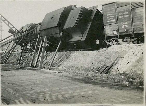 Schwarz-weiß Aufnahme: eine entgleiste Dampflok auf der linken Seite liegend, mit Holzbrettern vor dem weiteren Umkippen gestützt. Dahinter ein Anhänger stehend mit der Aufschrift "Deutsche Reichsbahn Kassel 25118"