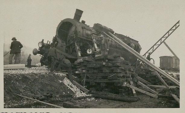Schwarz-weiß Aufnahme: eine entgleiste Dampflok auf der linken Seite liegend, von vorne seitlich fotografiert. Die Lok wird mit Holzbrettern gestützt.