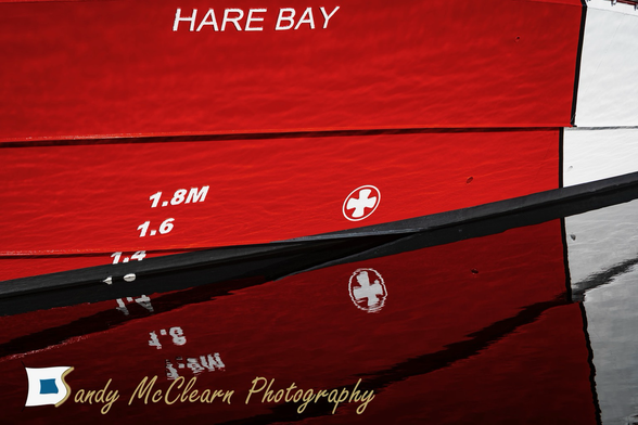 Hull of a red boat with a white stripe reflecting on calm water.