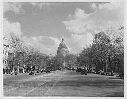 The image depicts a wide, open street leading towards the United States Capitol building with its iconic dome and Peace Monument at one end. The scene appears to be from an earlier era based on the style of vehicles visible in the distance - they look like early 20th-century automobiles. Trees line both sides of the road, their bare branches suggesting it might be late fall or winter.
The street is relatively empty with a few cars and horse-drawn carriages present, indicating that this image could have been taken during a time when personal automobile usage was not as prevalent as today. The sky above is partly cloudy but allows enough sunlight to illuminate the scene adequately.
Overall, it's an atmospheric black-and-white photograph capturing a moment in Washington D.C.'s past and showcasing one of its most famous landmarks.