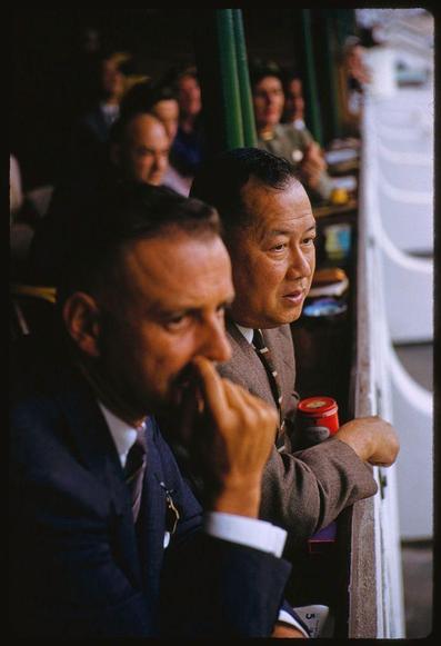 The photograph captures a moment in time during what appears to be an event or gathering. Two men are prominently featured, with one wearing a dark suit and tie, seemingly engrossed in thought as he rests his chin on his hand. The other man is dressed more casually in a brown jacket and holds a red can of drink by the window ledge where they seem seated.
In the background, several individuals appear to be engaged in various activities or conversations. Notably, there's someone wearing what looks like military-style clothing with insignia that might suggest an official capacity at this event.
The image has rich color saturation, particularly noticeable on the skin tones and darker shades of clothing. The composition suggests a candid snapshot rather than a posed photograph, capturing genuine reactions or interactions among those present. This was taken in Hong Kong as part of a Sports Illustrated assignment during July 1959, indicating its historical context within that timeframe.
The presence of Toni Frissell's name and connection to the magazine suggest she is known for her work across different fields including sports photography, highlighting this photograph’s significance beyond just being an ordinary moment captured. The date on which it was taken provides a direct link to understanding how life in Hong Kong during 1959 might have been depicted through media at that time.