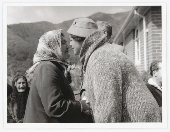 The black and white photograph depicts two women engaged in a close, affectionate interaction. The woman on the left is wearing a headscarf with patterns, possibly floral or paisley designs, draped over her shoulders, along with glasses resting atop her hat. She has dark hair visible under her cap and appears to be smiling as she looks towards the other woman.
The second woman is dressed in a textured coat that suggests it's cold weather. Her attention seems focused on something off-camera, possibly an object or another person not included within this frame. The two women are standing close together with their heads nearly touching, suggesting intimacy and connection between them.
In the background, there are other individuals who appear to be part of a larger group gathering outside what looks like a brick building, which could indicate some sort of communal event or meeting taking place in an outdoor rural setting given the visible hills or mountains in the distant landscape. The overall mood conveyed by this image is one of warmth and human connection despite potentially harsh weather conditions.