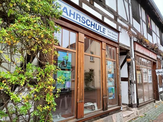 Fachwerkhaus mit Schaufenster, 'Fahrschule' Schriftzug, grüne Pflanzen, gepflasterter Weg, heller Tag.