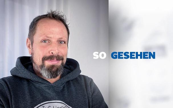 Mann mit Bart und dunkler Kapuzenjacke vor hellem Hintergrund, Logo auf der Jacke erkennbar.