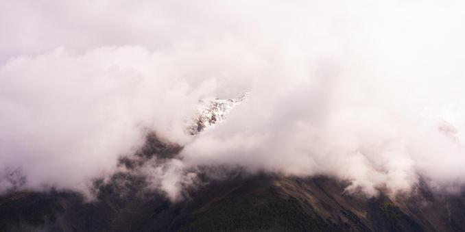 Cloud-covered mountain peak with sparse snow dusting visible through breaks in the clouds. Green-tinted slopes at the base, partially obscured by mist and fog. Soft, diffused light enhances the tranquil and atmospheric scene.