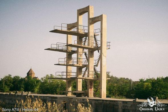 A dilapidated concrete diving platform stands tall, with a small domed building in the background. The platform has multiple levels and shows signs of graffiti.