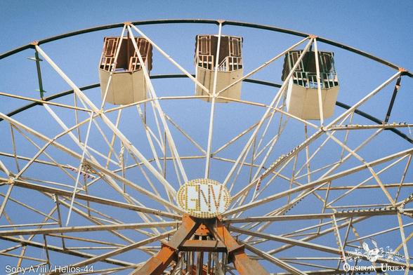 An old Ferris wheel with cream-colored carriages sits against a bright blue sky. The center displays the letters "GNV" in a golden oval.