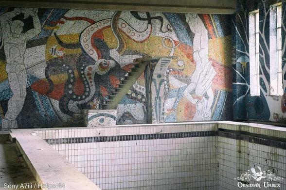 A mosaic mural depicts figures and marine life, over a vacant tiled pool.