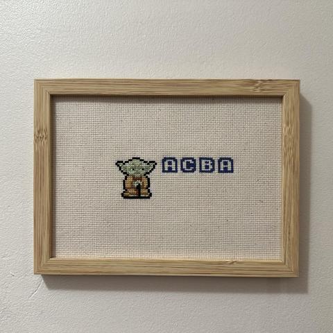 Yoda en pixel art avec les lettres ACBA dans une fonte de jeux videos. 
La broderie est sur une toile écrue, dans un cadre en bois.