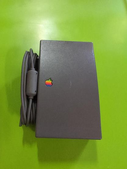 A dark gray plastic rectangle with a six-color Apple log inset on the body and a cord of the same color coiled against the left side of it. On a bright green background.