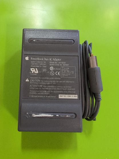 The underside of a dark gray plastic AC adaptor. The cord coiled next to it has a barrel connector. There are two rubber strips on the body of the adaptor. They both have a little paper stuck to them. Printed in white between the strips is an Apple logo and PowerBook Duo AC Adapter. Below that is company and technical info.