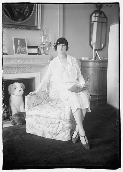 The image is a black-and-white portrait of a woman seated on an upholstered chair with a floral pattern. She is wearing a light-colored dress with a bow at the neckline and a pleated skirt. Her attire includes a dark hat and high-heeled shoes. The woman is holding an open book in her lap, and her posture is relaxed yet poised. A dog is sitting on the floor to the left of the chair, looking towards the camera. The background features a fireplace with a mantelpiece adorned with various decorative items, including a framed portrait, a chandelier, and a mirror. The overall setting suggests a formal or elegant interior, possibly a living room or a study. The photograph has a vintage quality, indicative of the early 20th century.