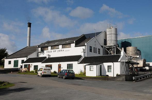 The Isle of Jura Distillery. The image shows a range of white industrial buildings stretching across the centre of the frame. The name of the distillery is on the side facing slightly to our left. There is a metal chimney on the left and a blue building on the right. The scene is in sunlight.