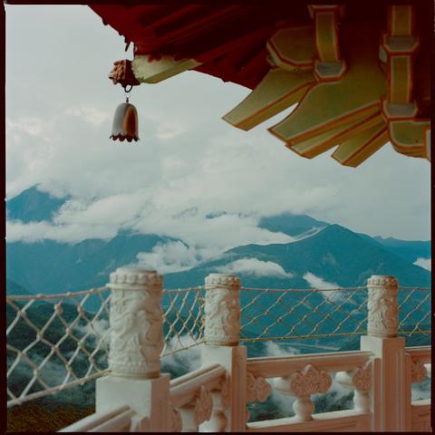 A traditional Chinese temple balcony with ornate white railings and a hanging bell under a red roof, overlooking mist covered mountains.