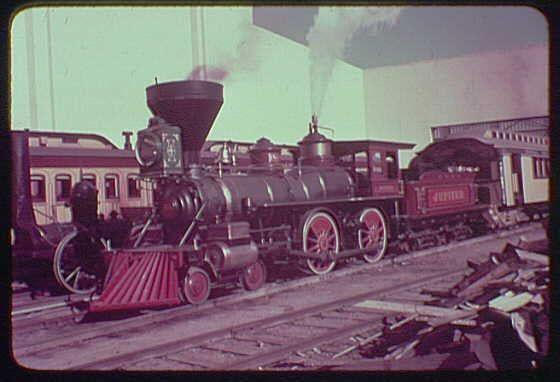 The image depicts a vintage steam locomotive, specifically labeled "Jupiter," at what appears to be an outdoor exhibition or fair. The Jupiter is painted in red with black trim and features prominent round wheels that are colored in shades of maroon and gold. It's situated on railway tracks, emitting smoke from its stack which suggests it's operational. In the background, there are various buildings, including a structure labeled "Pittsburgh" to one side.
The Jupiter locomotive is identified as part of an exhibit at the World's Fair held in 1939 or possibly around 1940 based on historical records and context provided by Gottscho-Schleisner Inc. The photograph captures not only the engine itself but also a glimpse into its era, showcasing how such locomotives were displayed for public engagement during that time period.
The image is in color with an overall warm tone, indicative of vintage photographs from early to mid-20th century exhibitions and fairs. It's worth noting that this photograph was taken by Gottscho-Schleisner Inc., a company known for its documentation of events such as the World's Fair during these years.
Overall, this image provides insight into how historical transportation equipment like steam locomotives were presented to the public in an exhibition setting and serves as an interesting piece of history regarding early 20th-century technological advancements.