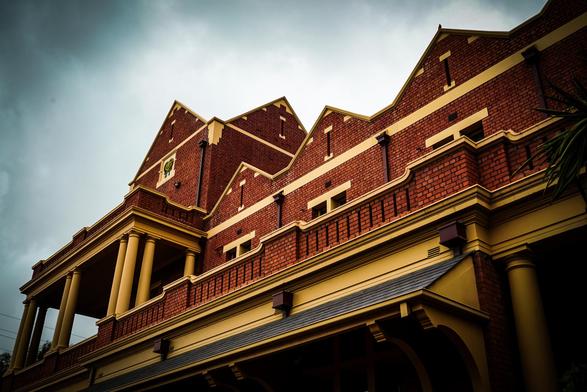 A low-angle, eye-level shot depicts the facade of a large, brick building under a cloudy, overcast sky. The building features a series of repeating, peaked roof sections along its length, each adorned with a beige decorative trim. A row of large, round, beige pillars support a partially visible second story balcony or porch area, which also has a beige trim. The brickwork is a deep reddish-brown color, contrasting with the lighter beige accents. The image appears to have been taken during the evening or on a dim day, creating strong shadows and highlighting the architectural details of the building.
Provided by @altbot, generated privately and locally using Gemma3:27b