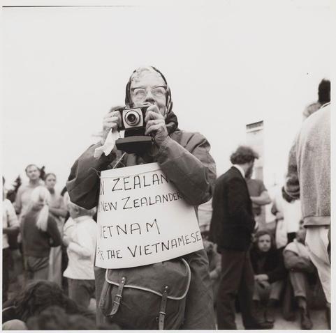 A black and white photograph features an individual taking a photo with their vintage camera. The person is wearing glasses, has grey hair tied back in a ponytail or bun, and appears to be older. They are holding up a piece of paper that reads "NEW ZEALANDER NEW ZEALANDER VIETNAM NEW ZEALANDER VET N AM FOR THE VIETNAMESE." The person is wearing glasses on their head, has a scarf around the neck area which could indicate cold weather or a fashion choice, and carries a messenger bag over one shoulder. In the background, there are various people in different states of engagement with each other; some appear to be talking while others look away from the camera. There's also an individual seated on what appears to be steps at the bottom right corner. The setting seems informal, potentially outdoors during daytime judging by the brightness and lighting.