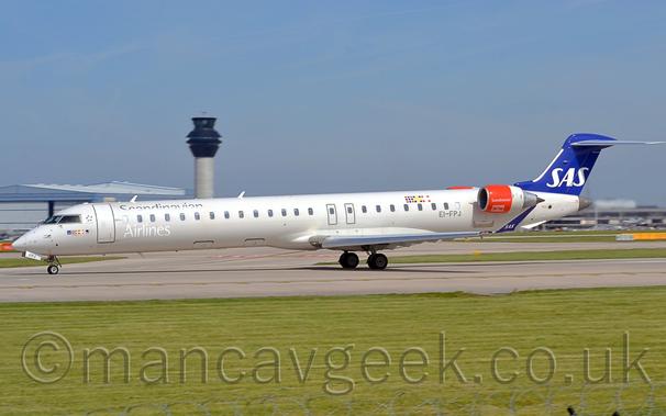 Side view of a long, thin, twin engined jet airliner with the engines mounted on the sides of the rear fuselage, parked on a runway waiting for it's take-off clearance.
The plane is largely white, with silvery-grey "Scandinavian Airlines" titles above and below the forward cabin windows, just aft of the forward door.
The grey registration "EI-FPJ" is on the side of the lower rear fuselage, just in front of the engine pods.
The engine pods themselves are red at the front and white at the rear, with white "Scandinavian" titles on the red portion.
The dark blue tail has white "SAS" titles at the bottom, a design repeated on the up-turned wingtips.
Green grass fills the foreground, lining the sides of the grey runway.
More grass lines taxiways in the background, with a grey terminal building in the distance on the right of the frame, with a large grey hangar and a tall grey and black tower on the left.
Blue sky with wisps of grey cloud fill the top of the frame.
