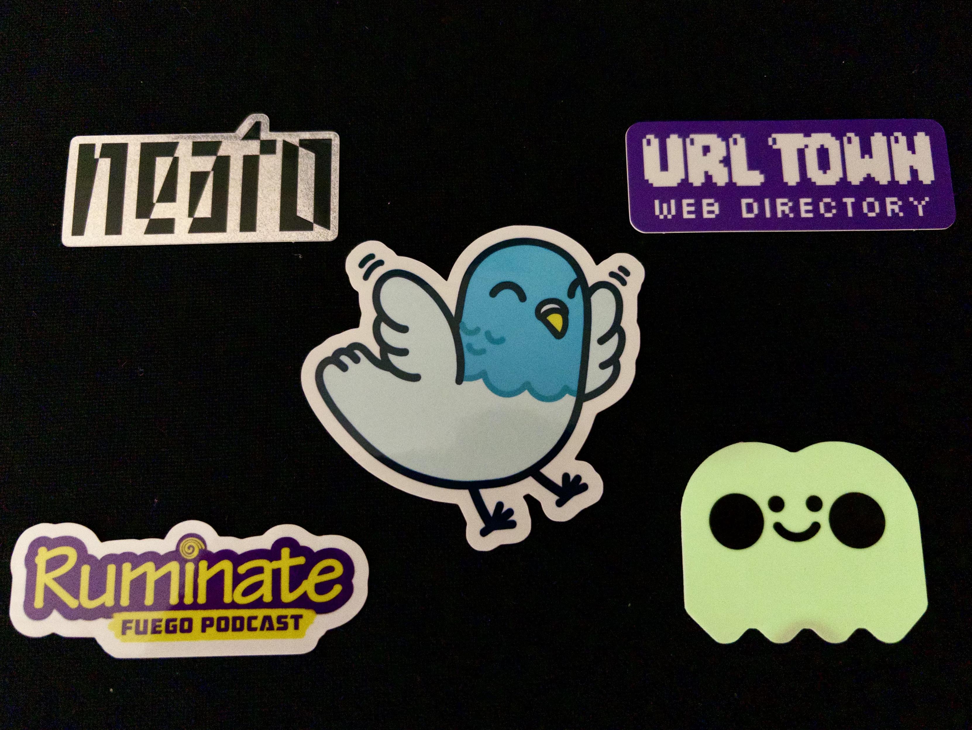 5 stickers arranged on a black surface: A glittery “neato” logo, a purple recatangle that says, “URL Town web directory,” a smiling cartoon pigeon with flapping wings, a purple and yellow logo for “Ruminate - fuego podcast,” a glow in the dark smiling ghost.