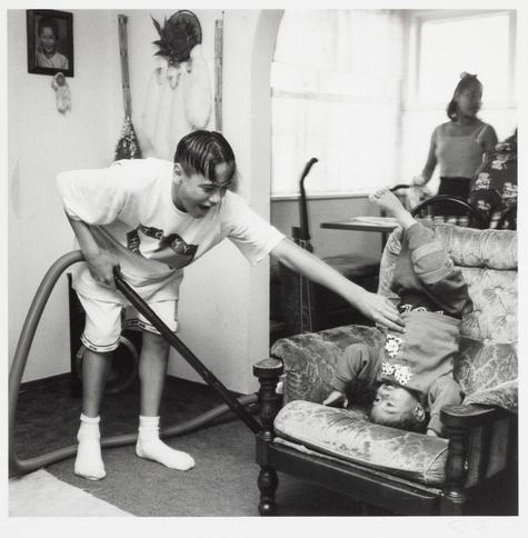 The black and white photograph depicts a domestic scene where an individual is using a vacuum cleaner on a child who appears to be lying back in a chair. The person operating the vacuum has short hair, wears shorts with rolled-up sleeves, socks, and holds the hose of the vacuum close to their body while laughing or expressing joy. Another figure can be seen through what seems like a glass door reflecting light, possibly outside, wearing loose-fitting clothing. In the background on shelves there are various items including what appear to be hats or ceremonial objects. The environment suggests this might be in a living room of an apartment or house. There's no direct indication of why the image is associated with Erua’s son from South Auckland based solely on its content, but it seems related as mentioned by way of caption reference.