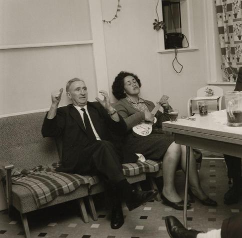 The image depicts an older Pakeha man with his hands raised and mouth open as if he is enthusiastically talking or singing, while sitting next to a Maori woman who appears relaxed. She has her eyes closed and seems to be enjoying herself, possibly dancing lightly on the sofa they are seated on.
They appear in front of a simple room setting decorated modestly with items like curtains, flowers above the window, and a telephone visible against the wall. There's another person partially seen at their right side who is not part of this interaction but seems to be sitting across from them possibly engaged in conversation or listening intently.
The environment suggests an informal gathering within a home setting during mid-20th-century times based on attire styles, furniture design, and black-and-white photography which was common for that era.