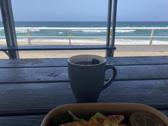Peppermint tea in a mug on a shaded picnic table with an ocean and beach view behind. Light sand, blues, greens and rows of white breaking waves. Part of a halloumi salad in a takeaway box in foreground.