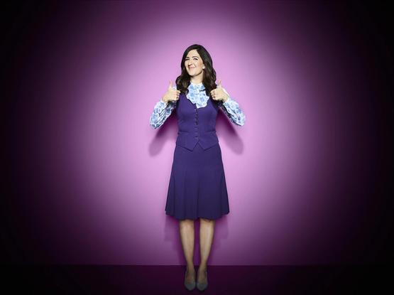 A woman in front of a lavender backdrop with a spotlight on her. She’s wearing a blue and white patterned shit and a dark purple dress she’s smiling and giving two thumbs up.