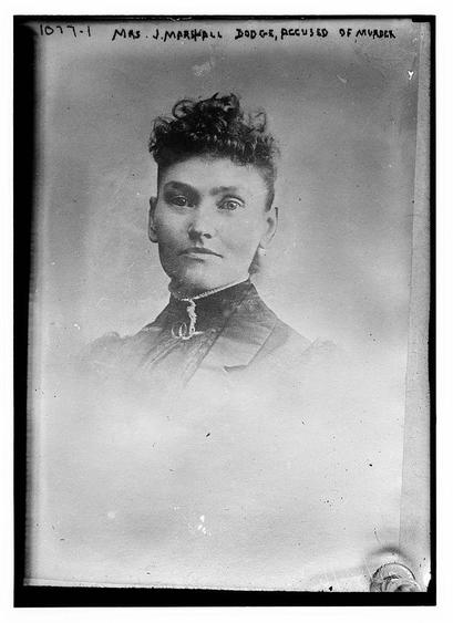 The image is a black-and-white photograph of a woman, likely from the late 19th or early 20th century, based on her attire and the style of the photograph. The woman has a serious expression and is looking directly at the camera. Her hair is styled in a short, curly fashion typical of the era. She is wearing a dark, formal dress with a high collar and a lace or embroidered detail around the neckline. The background of the photograph is plain and light-colored, which helps to focus attention on the subject. There is text at the top of the photograph that reads, "MRS. J. MARSHALL DODGE, ACCUSED OF MURDER," indicating that this photograph might be a mugshot or a formal portrait taken to document her involvement in a legal case. The photograph appears to be part of a collection, as indicated by the number "1077-1" at the top left corner.
