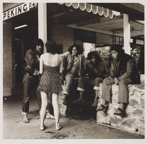The photograph captures a candid moment involving six individuals, likely from the 1970s or '80s judging by their fashion and hairstyles. A woman in a polka-dotted dress with her back to the camera stands as if she's about to walk away. She is facing four men seated on what appears to be a stone ledge outside an establishment named "Peking." The men are dressed casually, several wearing flared pants typical of that era and sandals or boots.
The scene conveys a sense of casual interaction in public space; the woman seems unaware or uninterested in their presence. Overhead signs indicate commercial establishments like "Goed Verkeer" (Good Traffic) and another with unclear text likely denoting food items, suggesting this might be near markets or eateries within Cuba Mall.
The black-and-white photo enhances its timeless quality, emphasizing textures and contrasts without the distraction of color. No discernible biases are present in describing these individuals; they appear as everyday people engaging in life's simple moments captured through a lens with artistic intent by Ans Westra.