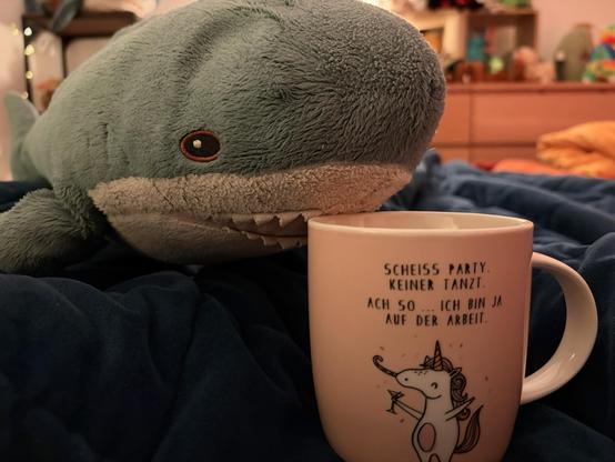 English:A plush Blåhaj is lying on a blue blanket, next to a mug with a humorous text in German. The mug features a cartoon unicorn holding a drink. The background hints at a cozy, colorful room.

Deutsch: Ein Plüsch-Blåhaj liegt auf einer blauen Decke neben einer Tasse mit einem humorvollen Text auf Deutsch. Der Becher zeigt ein Cartoon-Einhorn, das ein Getränk hält. Der Hintergrund deutet auf ein gemütliches, farbenfrohes Zimmer hin.